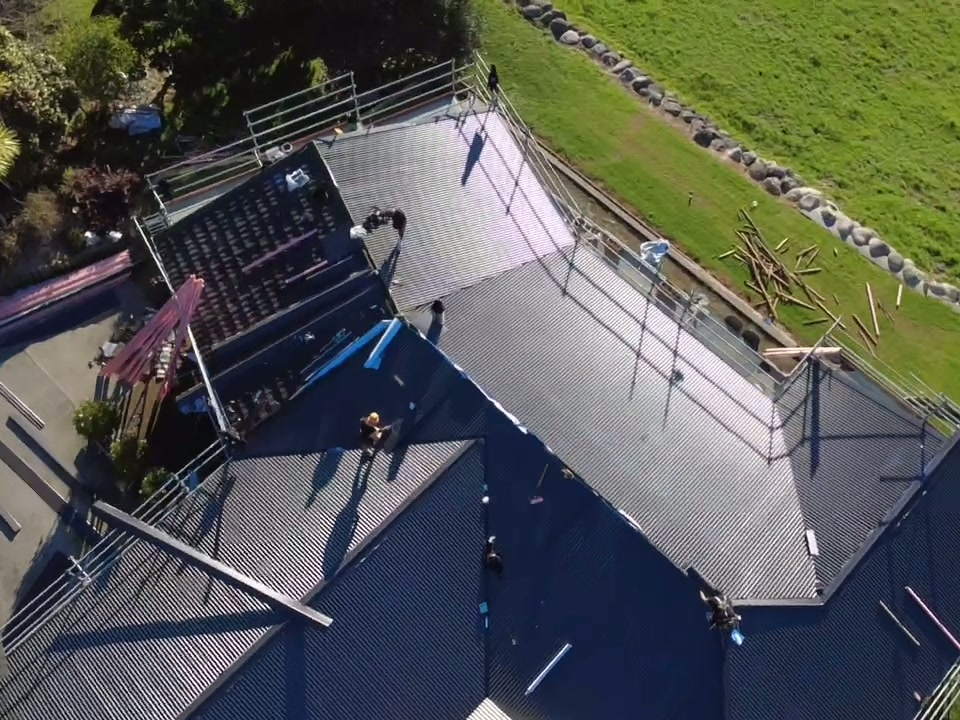 Two roofers laying dark longrun sheets on a hip roof, straight down from the drone