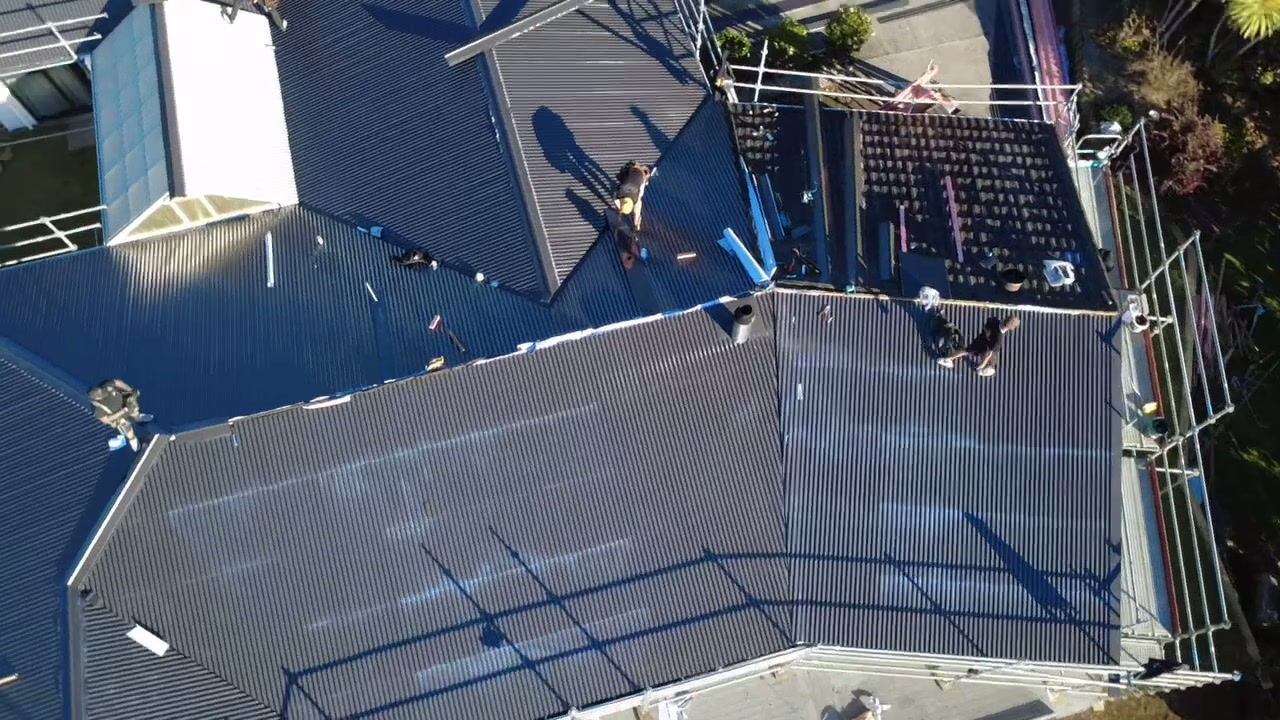 New dark longrun roofing laid across a Taranaki house with a roofer at the ridge and scaffold around the edge