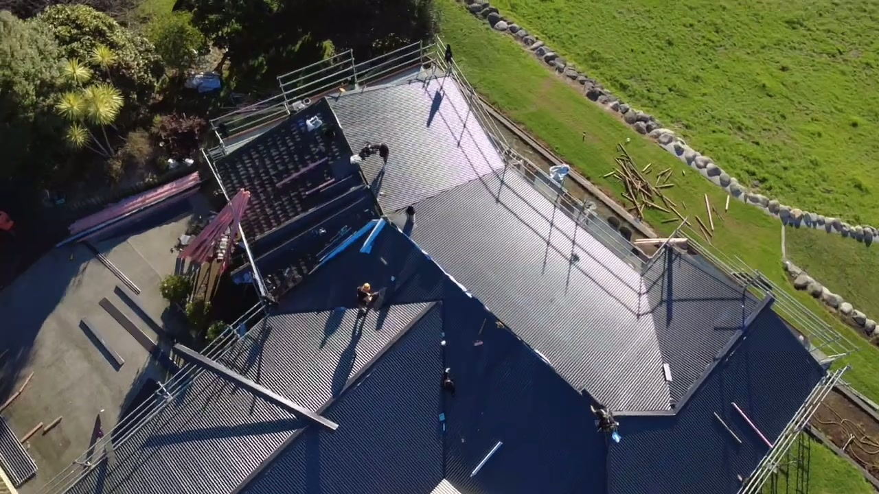Two roofers laying new dark longrun sheets on a hip roof, seen from directly above