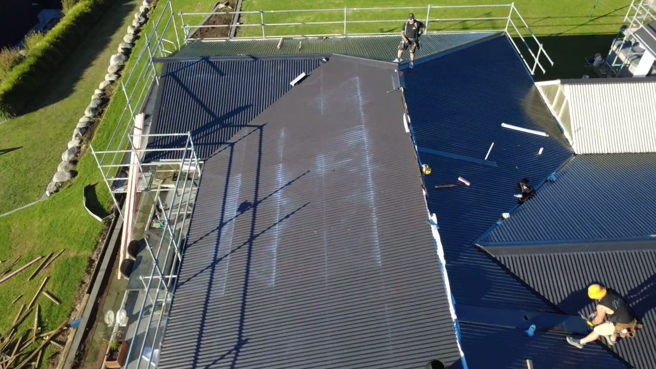Roofer working along a long run of new dark corrugate roofing with the scaffold rail in the foreground